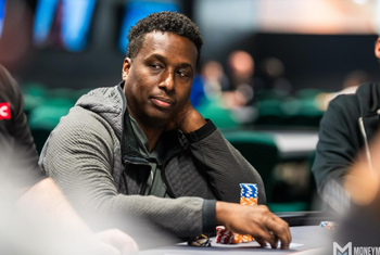 A person sitting at a poker table, looking to the side with a thoughtful expression. They are wearing a gray jacket and have a stack of poker chips in front of them. The background is blurred, showing other tables and players.