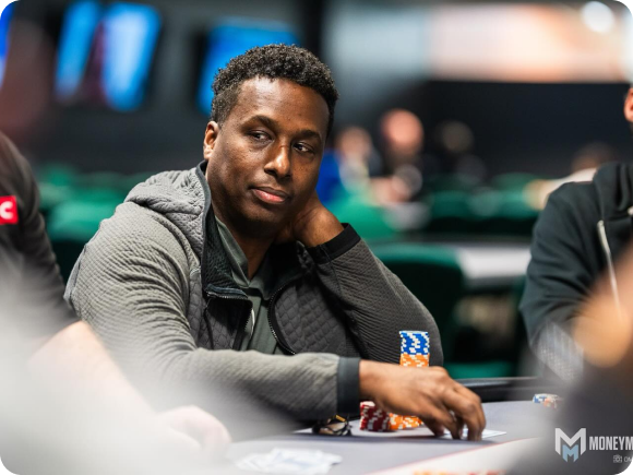 A person sitting at a poker table, looking to the side with a thoughtful expression. They are wearing a gray jacket and have a stack of poker chips in front of them. The background is blurred, showing other tables and players.